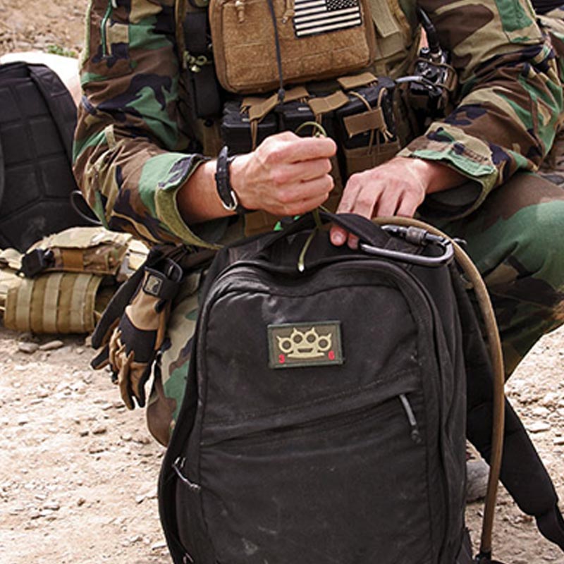 Person in camouflage military gear kneeling and holding a black backpack with a patch on a rocky surface.