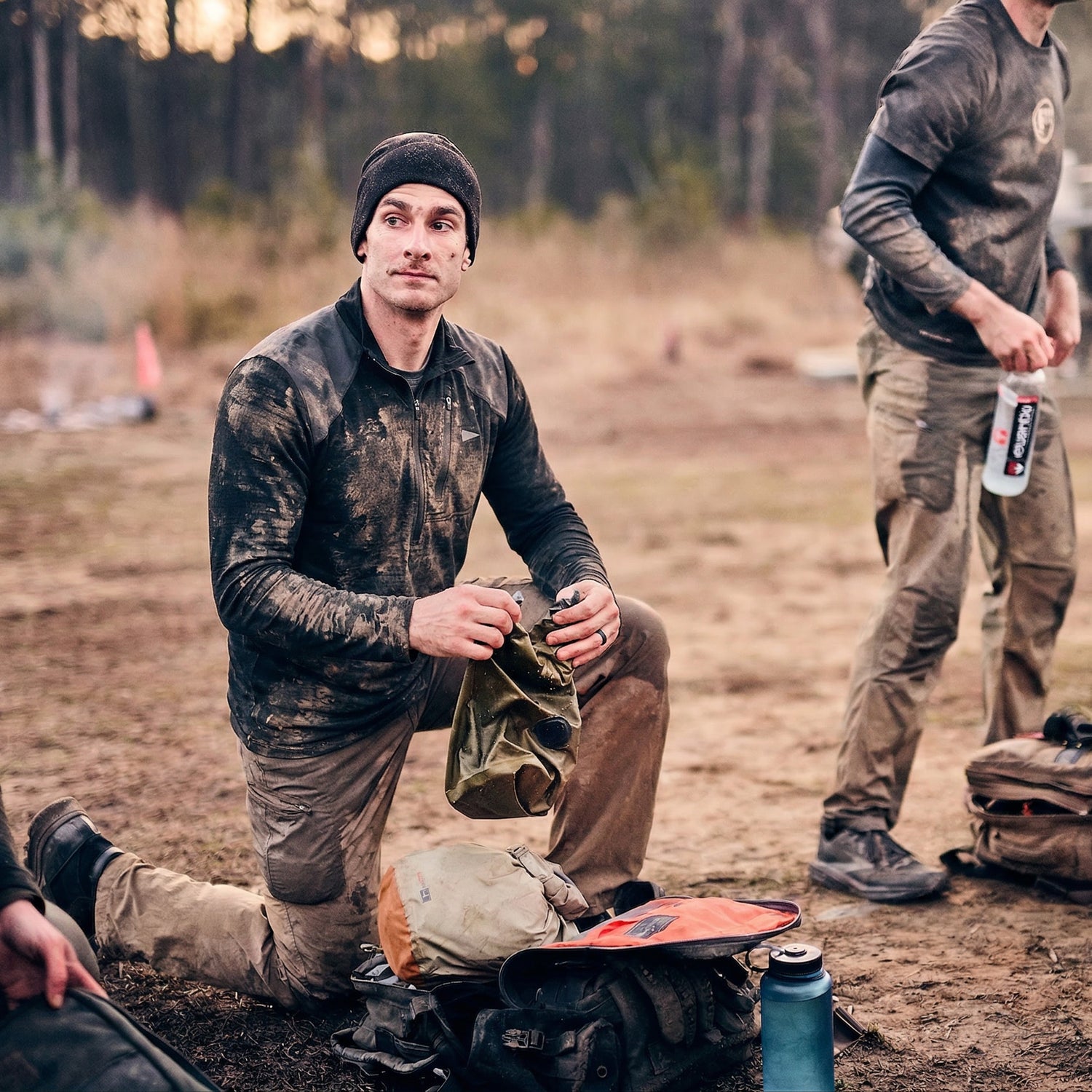 A man kneels on muddy ground holding a cap, while another stands nearby; both wear dirty outdoor gear, including the Men’s Commando Half Zip - Merino Wool sweater with reinforced shoulders for extra durability.