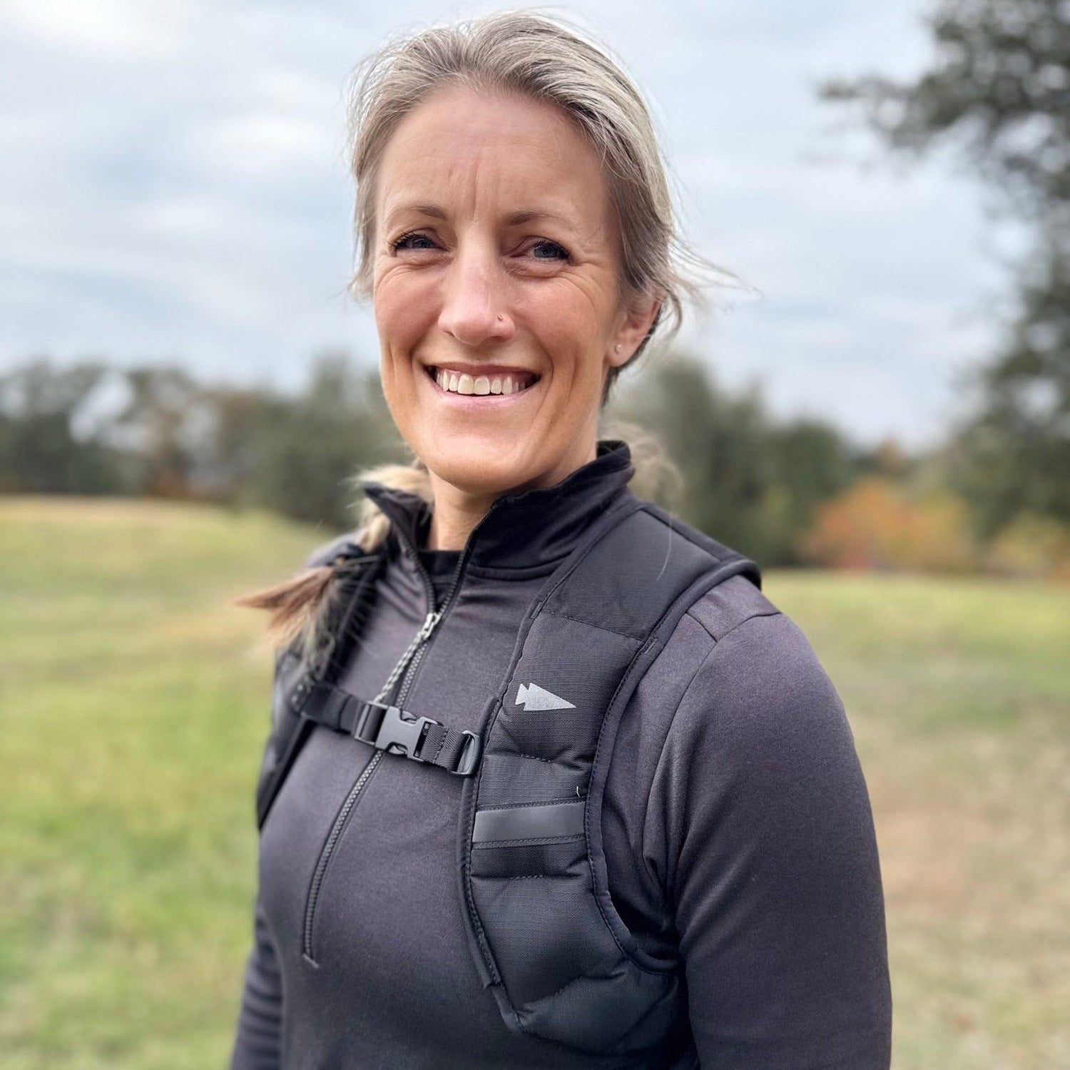 Smiling woman in a black jacket and backpack outdoors with trees and grass in the background.