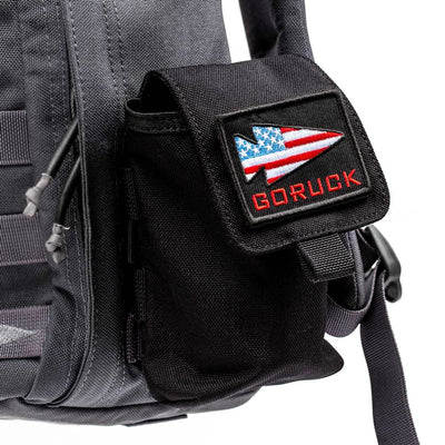 A black Simple Side Pocket by GORUCK is attached to a gray backpack, featuring an American flag arrow design patch. The setup includes patch-friendly VELCRO and the GORUCK logo in red, making it perfect for custom flair.
