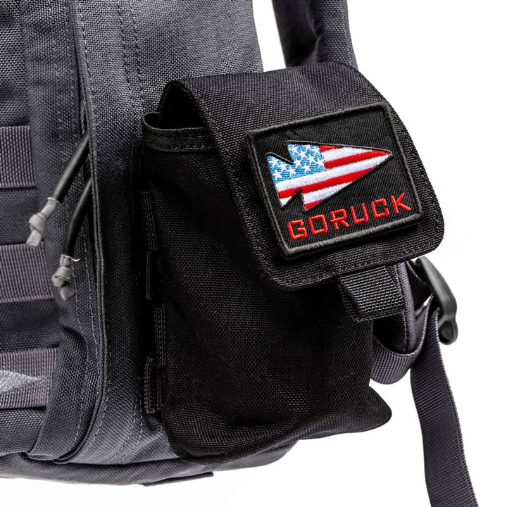A black Simple Side Pocket by GORUCK is attached to a gray backpack, featuring an American flag arrow design patch. The setup includes patch-friendly VELCRO and the GORUCK logo in red, making it perfect for custom flair.