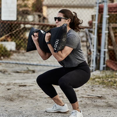 Wearing sunglasses, a person is exercising outdoors with the 100-pound Simple Training Sandbags by GORUCK, which features padded handles. They are squatting with a chain-link fence and some outdoor equipment visible in the background.