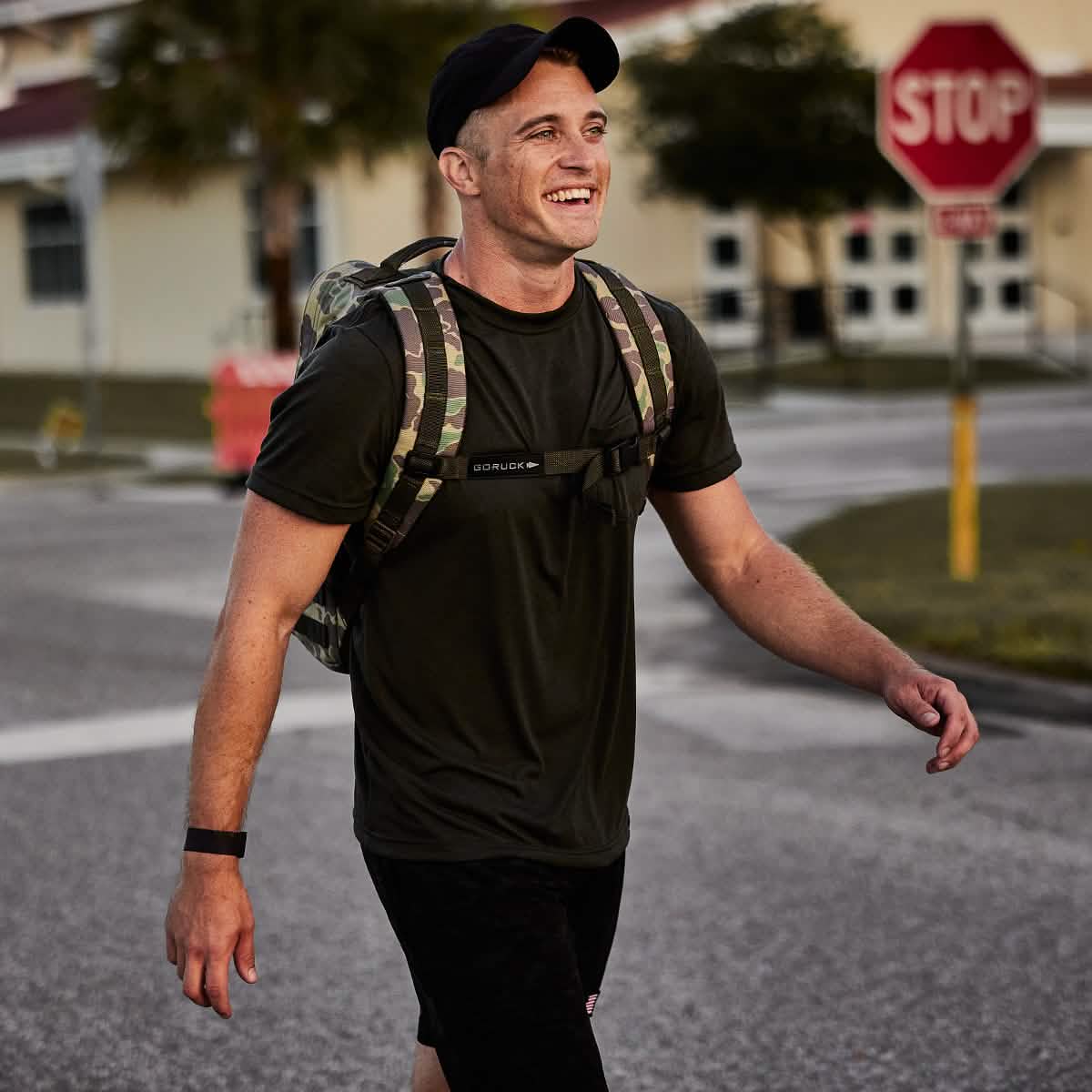 A person wearing a dark sweat-wicking t-shirt, a GORUCK Performance TAC Hat - Slick - TOUGHDRY, and a camouflage backpack strolls down the street, smiling. A stop sign and buildings are visible in the background. The scene looks casual and the weather appears pleasant.