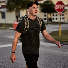 A person wearing a dark sweat-wicking t-shirt, a GORUCK Performance TAC Hat - Slick - TOUGHDRY, and a camouflage backpack strolls down the street, smiling. A stop sign and buildings are visible in the background. The scene looks casual and the weather appears pleasant.