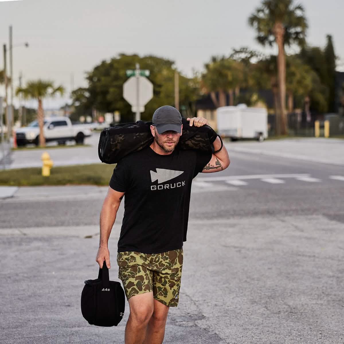 A man in a GORUCK Performance TAC Hat - Slick - TOUGHDRY, carries a large bag on his shoulder and a smaller one in his other hand. He sports a logoed T-shirt made of sweat-wicking ToughDry® fabric along with camo shorts, as he strolls along a street lined with palm trees and vehicles.