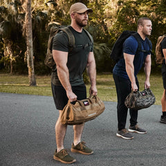 Two men in workout gear are lifting weighted bags during an outdoor training session on a paved path, surrounded by trees and grass. One is wearing a GORUCK Performance TAC Hat - Slick made from TOUGHDRY material for added comfort, while both have backpacks with them. The scene takes place during daylight.