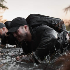 Wearing a GORUCK Performance TAC Hat - Slick - TOUGHDRY and a sturdy backpack, a person is crawling through muddy water with focus and determination. The outdoor setting includes another individual partially visible nearby, set against an overcast sky.