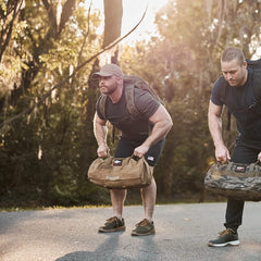 Two men exercise outdoors on a paved path in a forest, their GORUCK Performance TAC Hats with TOUGHDRY technology combined with their sweat-wicking T-shirts and shorts making for the perfect workout gear. Bent forward with weighted bags, sunlight filters through the trees as they move in sync, sneakers pounding rhythmically against the trail.