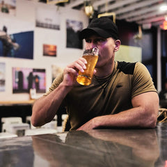 Someone sporting a sleek black Performance TAC Hat - Slick by GORUCK and an olive shirt is seated at a bar, enjoying a pint of beer. Framed photos adorn the background, contributing to the cozy atmosphere with soft lighting that beautifully complements the hat's Sweat-wicking TOUGHDRY® fabric.