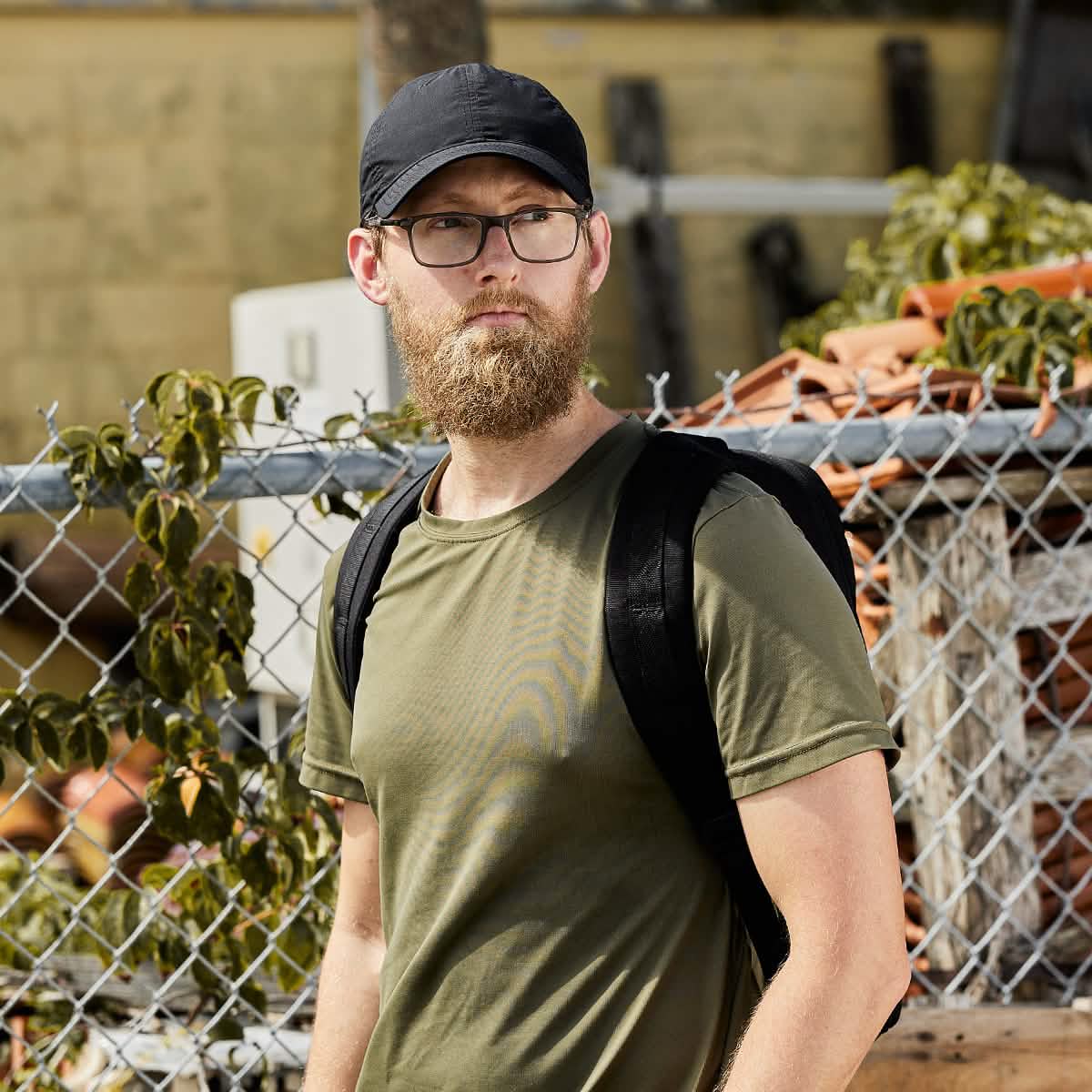 A man sporting a beard and glasses, wearing a black GORUCK Performance TAC Hat - Slick made from Sweat-wicking TOUGHDRY fabric, stands in front of a chain-link fence. He's dressed in an olive green T-shirt and is carrying a black backpack. The background is filled with plants and a stack of bricks.