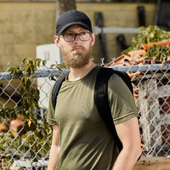 A man sporting a beard and glasses, wearing a black GORUCK Performance TAC Hat - Slick made from Sweat-wicking TOUGHDRY fabric, stands in front of a chain-link fence. He's dressed in an olive green T-shirt and is carrying a black backpack. The background is filled with plants and a stack of bricks.