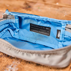 Open gray Shadow Pocket with a blue interior, GORUCK label, and zipper on a wooden surface—ideal for organizing your rucksack. Made of durable 500D Cordura, Shadow Pockets keep your essentials secure and easy to access.
