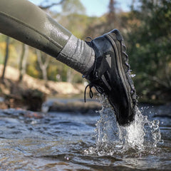 A person wearing waterproof shoes steps into a shallow, flowing stream, causing a splash. The scene is outdoors with blurred greenery in the background. The focus is on the shoe and water where the GORUCK Merino Challenge Socks - Trail Cuff offer snug protection.