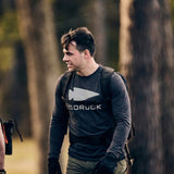 A man in the Men's GORUCK Spearhead Long Sleeve - Tri-BlendX, featuring antimicrobial treatment, smiles outdoors with a backpack in a wooded area.
