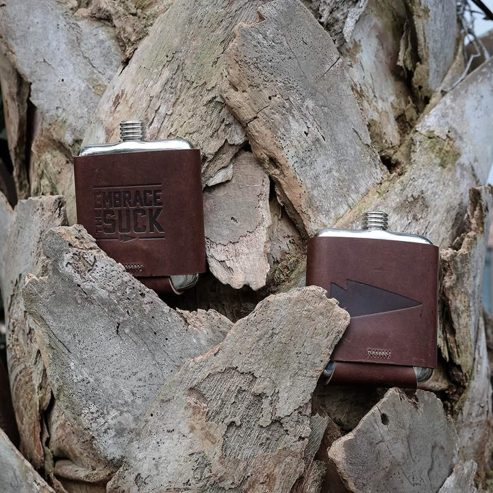 Resting on the textured trunk of a palm tree are two small, GORUCK Leather Wrapped Flasks by Built By Scars. Both flasks feature metal tops; one is crafted from stainless steel and embossed with "EMBRACE THE SUCK," while the other sports a minimalist arrow design, making them perfect companions for any adventure.