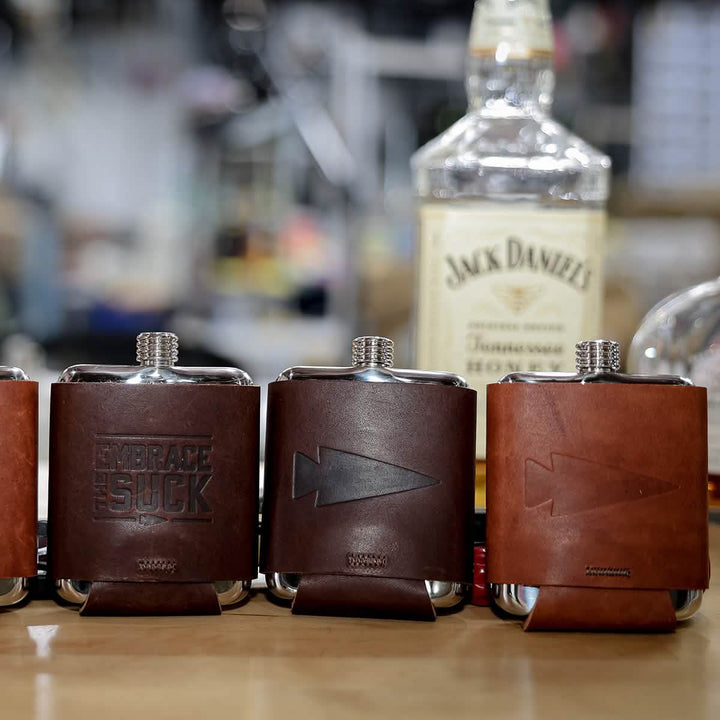Three GORUCK Built By Scars leather-wrapped hip flasks, featuring embossed designs on stainless steel, are displayed on a wooden surface. A bottle of Jack Daniels is visible in the background, creating an adventurous atmosphere indoors—adventure in every pour.
