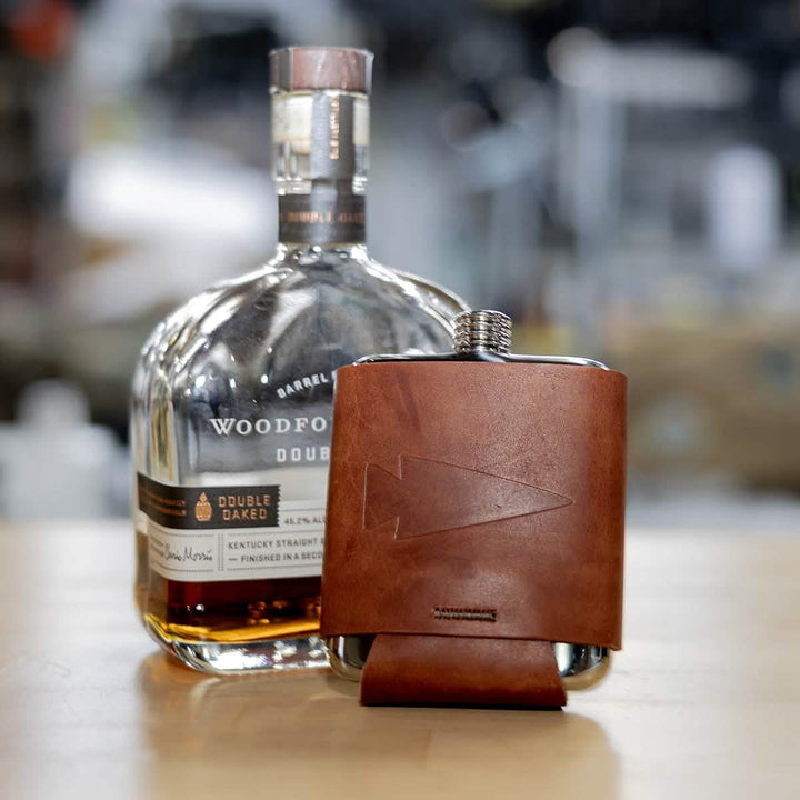 A bottle of Woodford Reserve Double Oaked bourbon sits on a wooden table, promising an adventure in every pour. Beside it lies the Leather Wrapped Flask by GORUCK, featuring a stainless steel body wrapped in brown leather with an arrow design, while the background remains blurred.