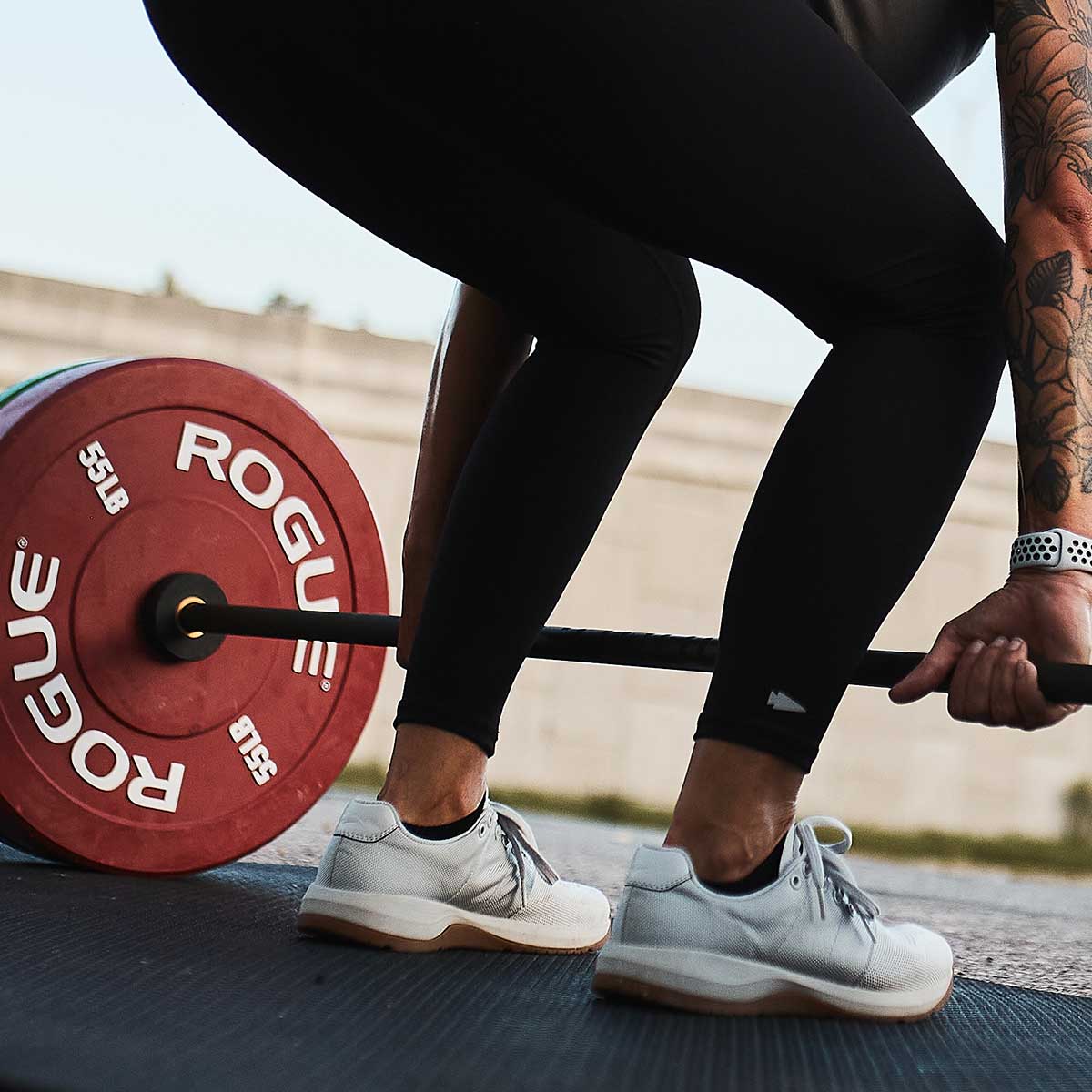 Person in athletic wear preparing to lift a loaded barbell with red Rogue weight plates outdoors.