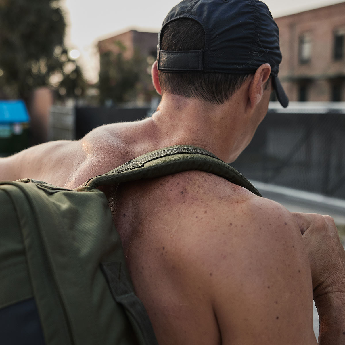 Shirtless man wearing a cap carries a backpack outdoors on a sunny day.