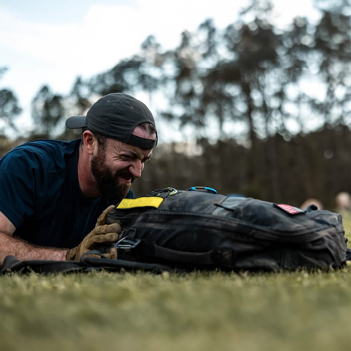 A man in a GORUCK Performance TAC Hat - Slick - TOUGHDRY crouches on the grass, smiling at a black backpack. His gloves and dark shirt enhance the scene, while trees and a cloudy sky create an ideal backdrop for adventure, with sweat-wicking gear keeping him comfortable all day long.