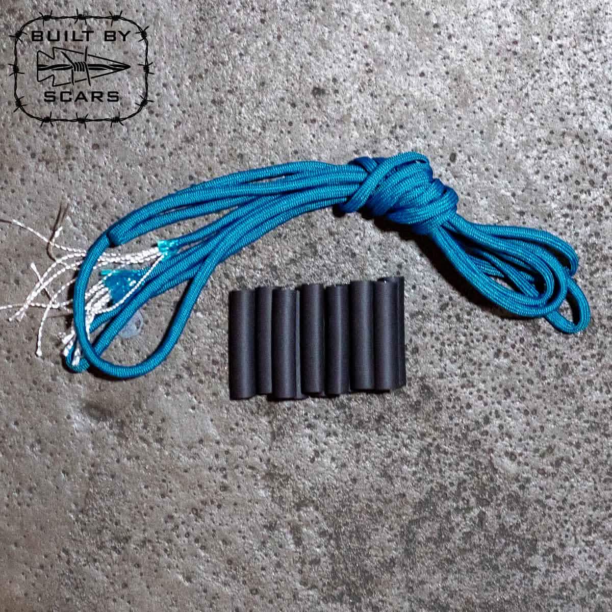 A coiled blue paracord from the GORUCK Paracord Kit - Built By SCARS lies alongside six sleek black heat shrink tubes on a textured gray surface. In the top left corner, there's a logo featuring a stylized fish with the words 