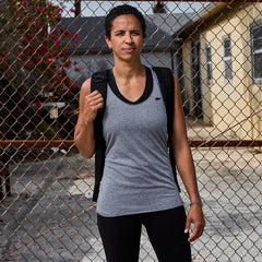 Woman wearing grey and black sleeveless athletic shirt with small logo, carrying black backpack, standing by rusty chain-link fence
