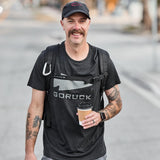 Man wearing black GORUCK tee, backpack, and hat, holding coffee on urban street