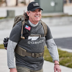 Man wearing GORUCK The Three Rules charcoal tee, tactical backpack, and black cap with American flag patch