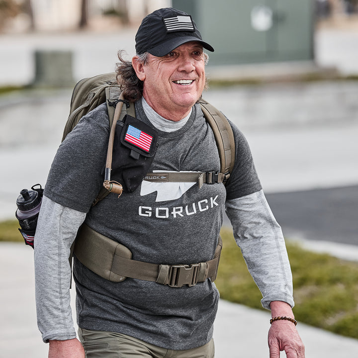 A smiling man outdoors wears the GORUCK Three Rules Tee - Tri-Blend and cap, carrying a backpack with an American flag patch—ideal for standout photos.
