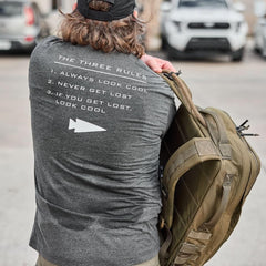 Man wearing charcoal GORUCK The Three Rules tee with white text and carrying a tan tactical rucksack outdoors