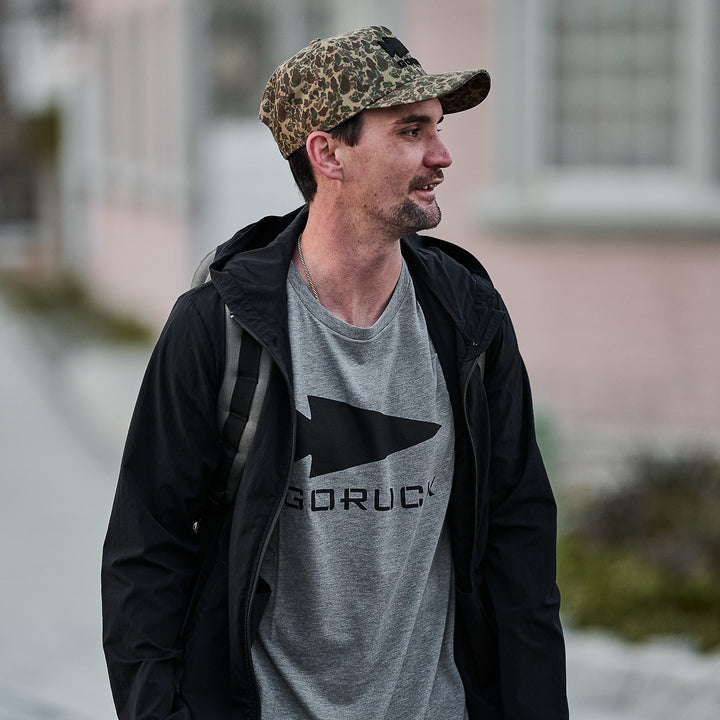 Wearing a camo hat, black jacket, and the Three Rules Tee - Tri-Blend, this semi-famous adventurer walks outdoors with a backpack—perfect for candid adventure photos.