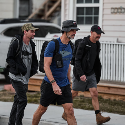 Three men wearing Three Rules Tee - Tri-Blend casual shirts and hats walk together on a sidewalk in front of a house, as if posing for candid photos.