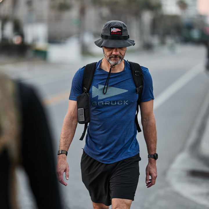A semi-famous man wears the Three Rules Tee - Tri-Blend in blue, black shorts, and a hat as he walks outdoors with a backpack and water tube in his mouth.