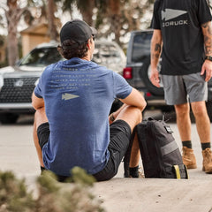 Man wearing GORUCK navy 'The Three Rules' t-shirt sitting outdoors with black GORUCK backpack in urban setting