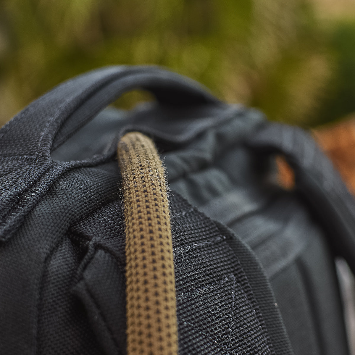Close-up of a black backpack with a tan cord looped over the handle, blurred green background.