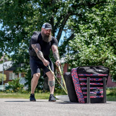 Bearded man with tattoos pulling a GORUCK weighted sled outdoors on pavement, surrounded by trees and houses