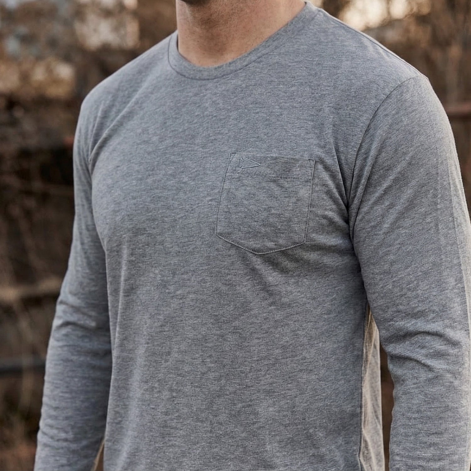 A man wears the Men's Long Sleeve Pocket Tee - Tri-BlendX, featuring a chest pocket and made from premium, durable Tri-BlendX fabric, while standing outdoors.