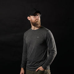 A man with a beard, wearing glasses, a black cap, and the Men's Long Sleeve Pocket Tee - Tri-BlendX in dark color stands against a black background.