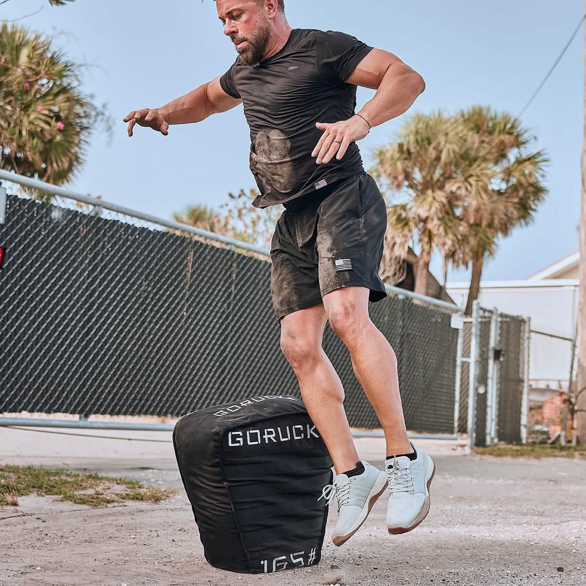 A man in athletic wear, wearing the Men’s USA Performance Tee - ToughMesh, jumps onto a large weighted sandbag outdoors near a fenced area with palm trees.