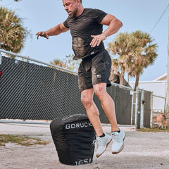 Athletic man jumping over a black GORUCK sandbag outdoors in casual workout gear near a fence and palm trees