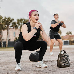 Two people perform squats outdoors. The woman in front, sporting pink hair, wears the GORUCK Training Weight Vest 2.0 in black. Her companion in the background dons a gray outfit. Nearby rest a weighted bag and ball, with palm trees and buildings serving as the backdrop.