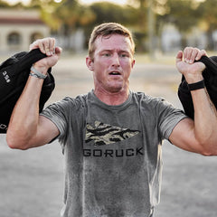 A person exercises outdoors with a pair of Sand Kettlebells on their shoulders, dressed in a gray GORUCK T-shirt. The blurred background highlights their movement and focus on the workout.