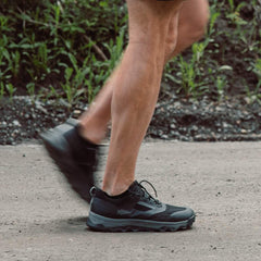 Close-up of a person wearing black GORUCK tactical shoes walking on a paved path with greenery in the background