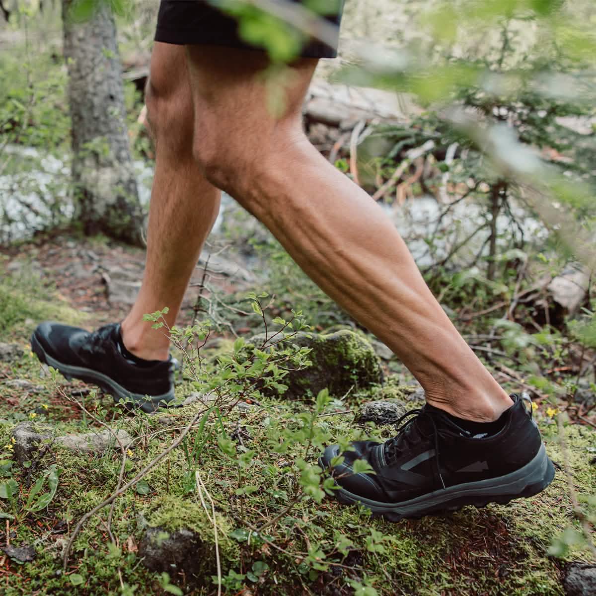 A person wearing the Mackall - Black + Forged Iron trail shoes and shorts from GORUCK is walking briskly on a moss-covered forest path, evoking the feel of Special Forces training. They're surrounded by lush greenery as a small creek babbles gently in the background.