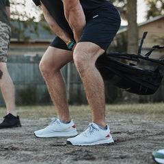 Wearing the Men's Rough Runner - Light Grey + Red, featuring a Gradient Density EVA midsole, a person in shorts lifts a sandbag outdoors to prep for squats.
