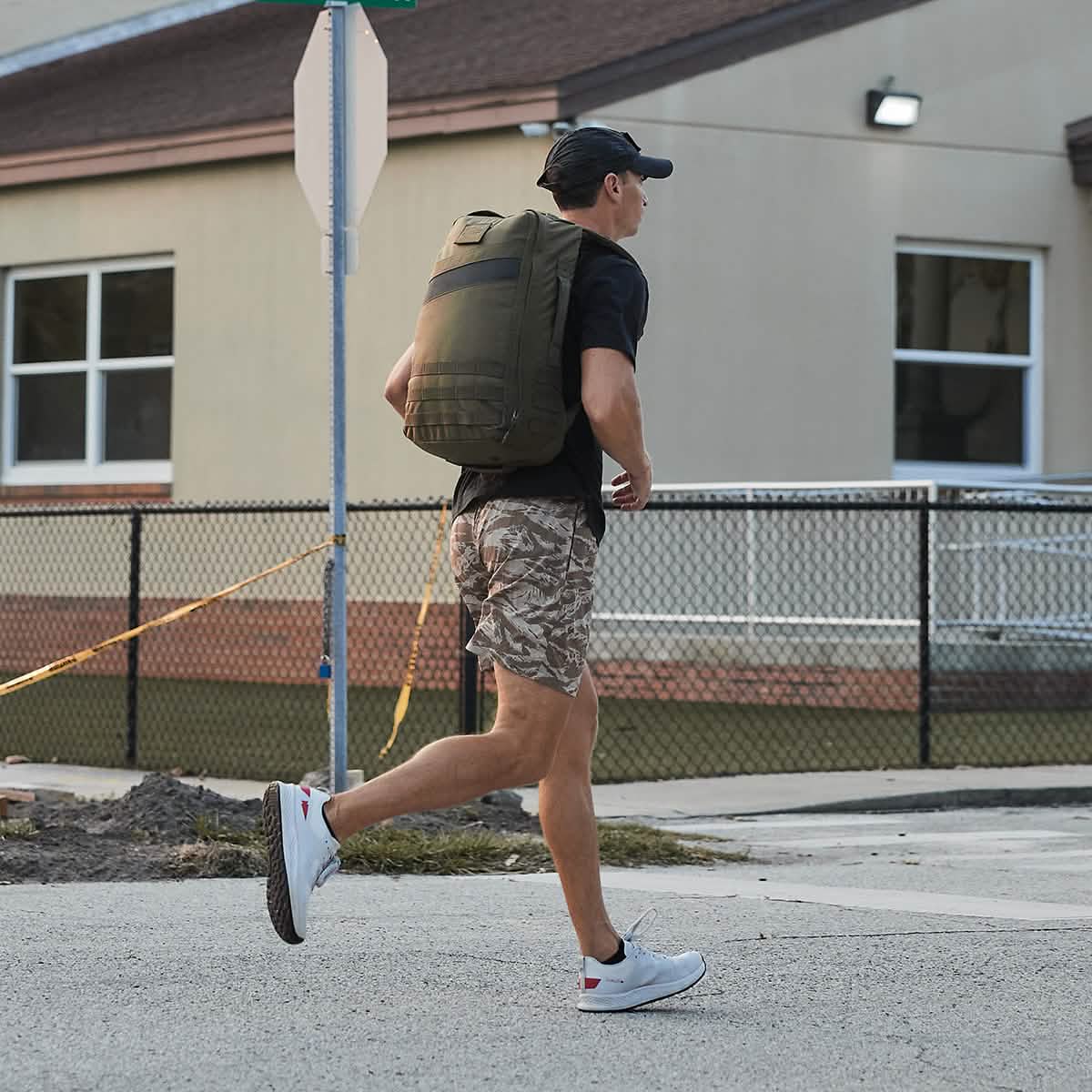 A man wearing a cap, shorts, and Men's Rough Runner - Light Grey + Red shoes walks outside with a large backpack near a building and fence.