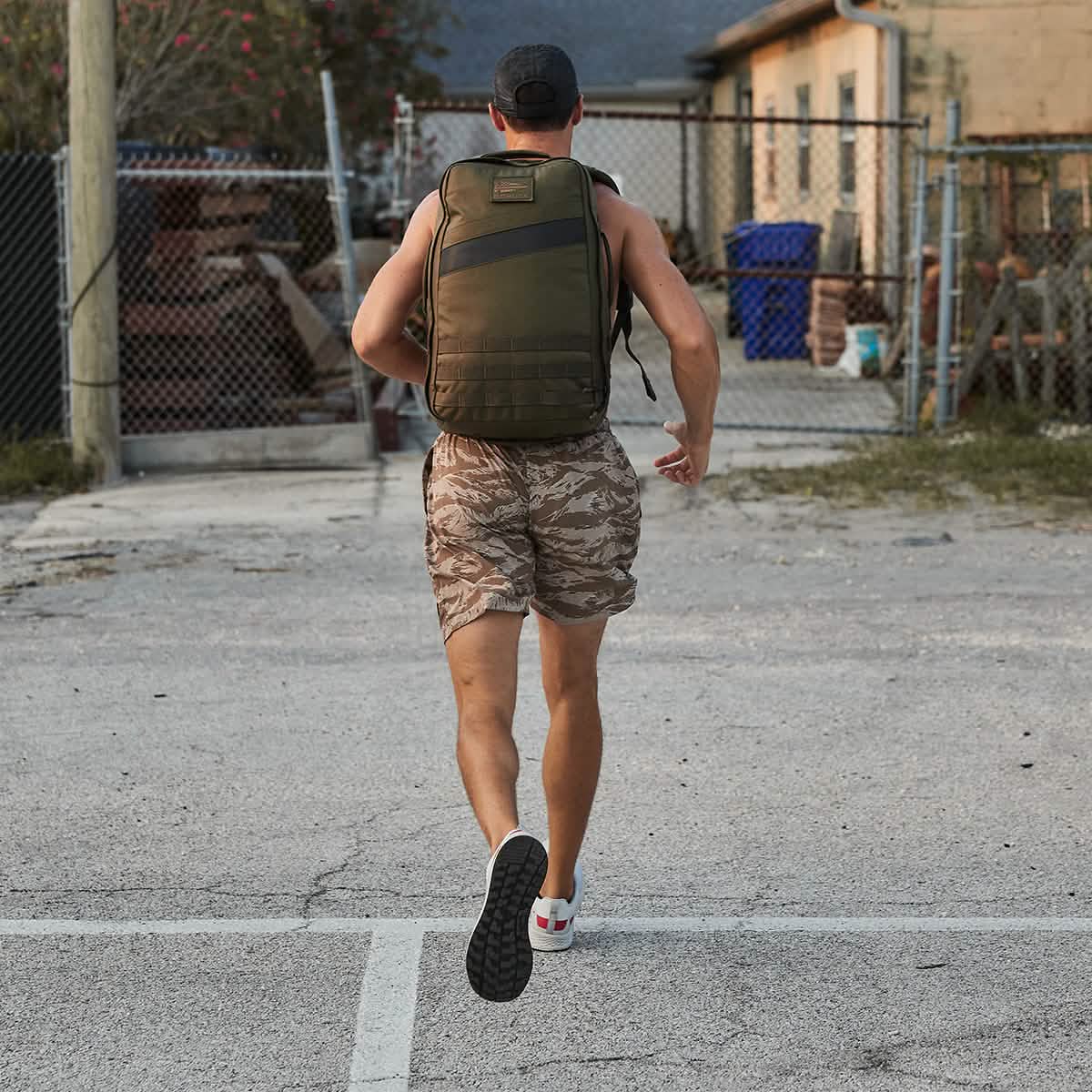 A person in camo shorts runs outdoors with a green backpack, seen from behind, wearing Men's Rough Runner - Light Grey + Red shoes for comfort on the trail.