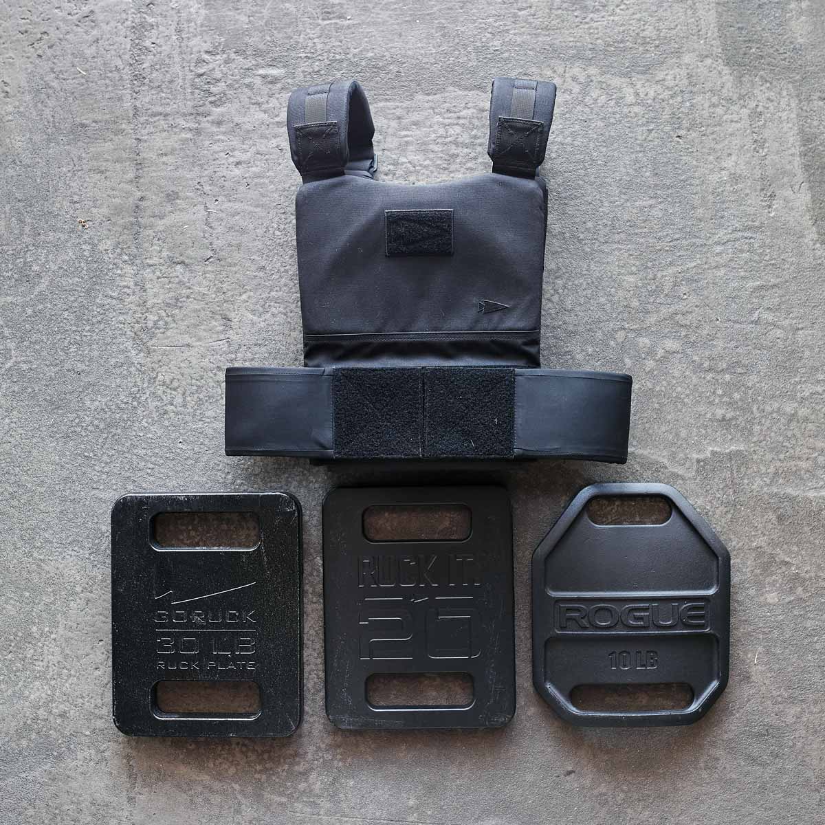 Black weighted vest and three different weight plates on a gray concrete surface.