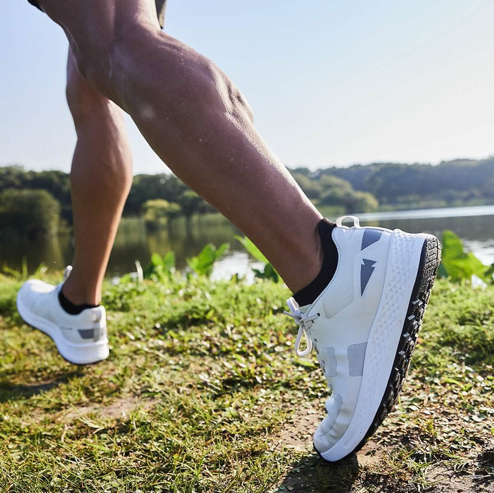 A person jogging outdoors next to a lake embodies the spirit of GORUCK's Men's Rough Runner shoes in Light Grey + Dark Grey. The focus is on their legs and athletic footwear, set against a backdrop of green grass and trees under a clear blue sky.