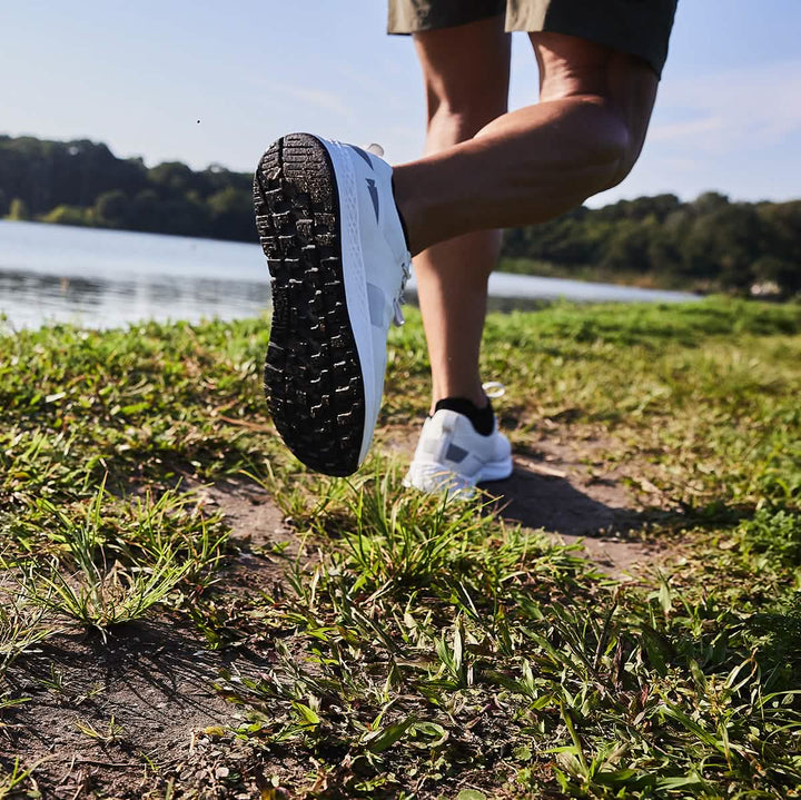 A person walking along a grassy path by a lake, wearing the Men's Rough Runner in Light Grey and Dark Grey from GORUCK. The focus is on the sole of the shoe as the foot is lifted, with green trees and a blue sky in the background.